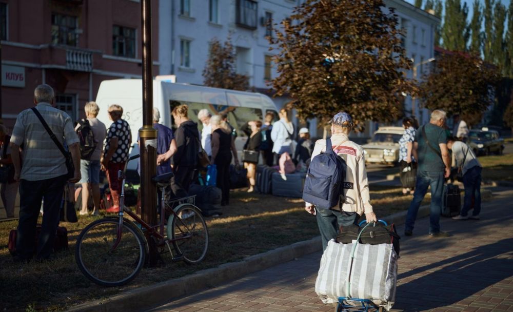 Люди з валізами, сумками та візком очікують біля мікроавтобуса на міській вулиці.