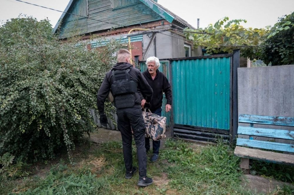 Волонтер у бронежилеті допомагає літньому чоловікові винести з двору сумку.